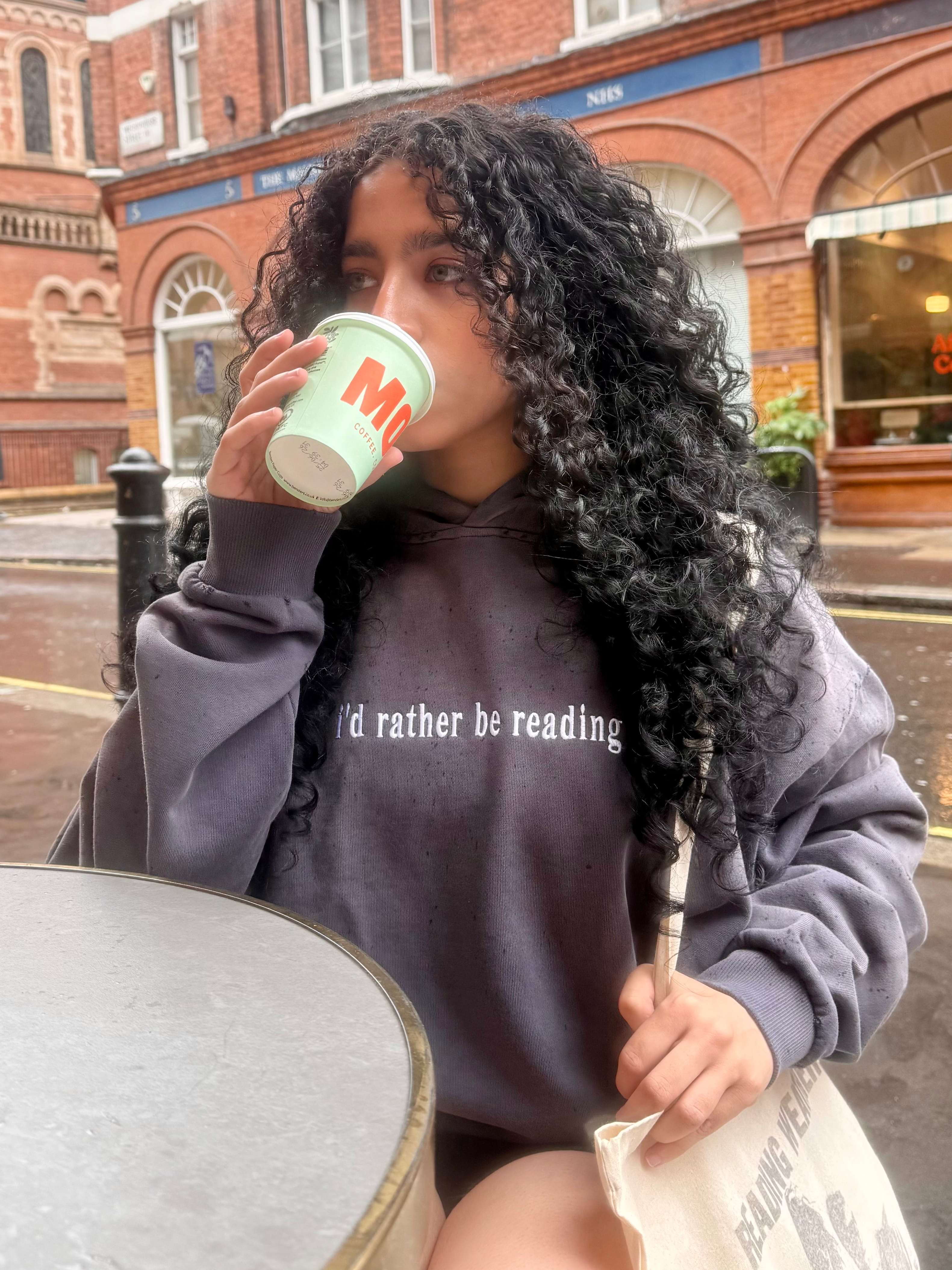 A woman with long, curly black hair sits at an outdoor cafe table in front of a brick building, wearing a dark gray hoodie with the text 'i'd rather be reading' and holding a light green paper cup with red text.