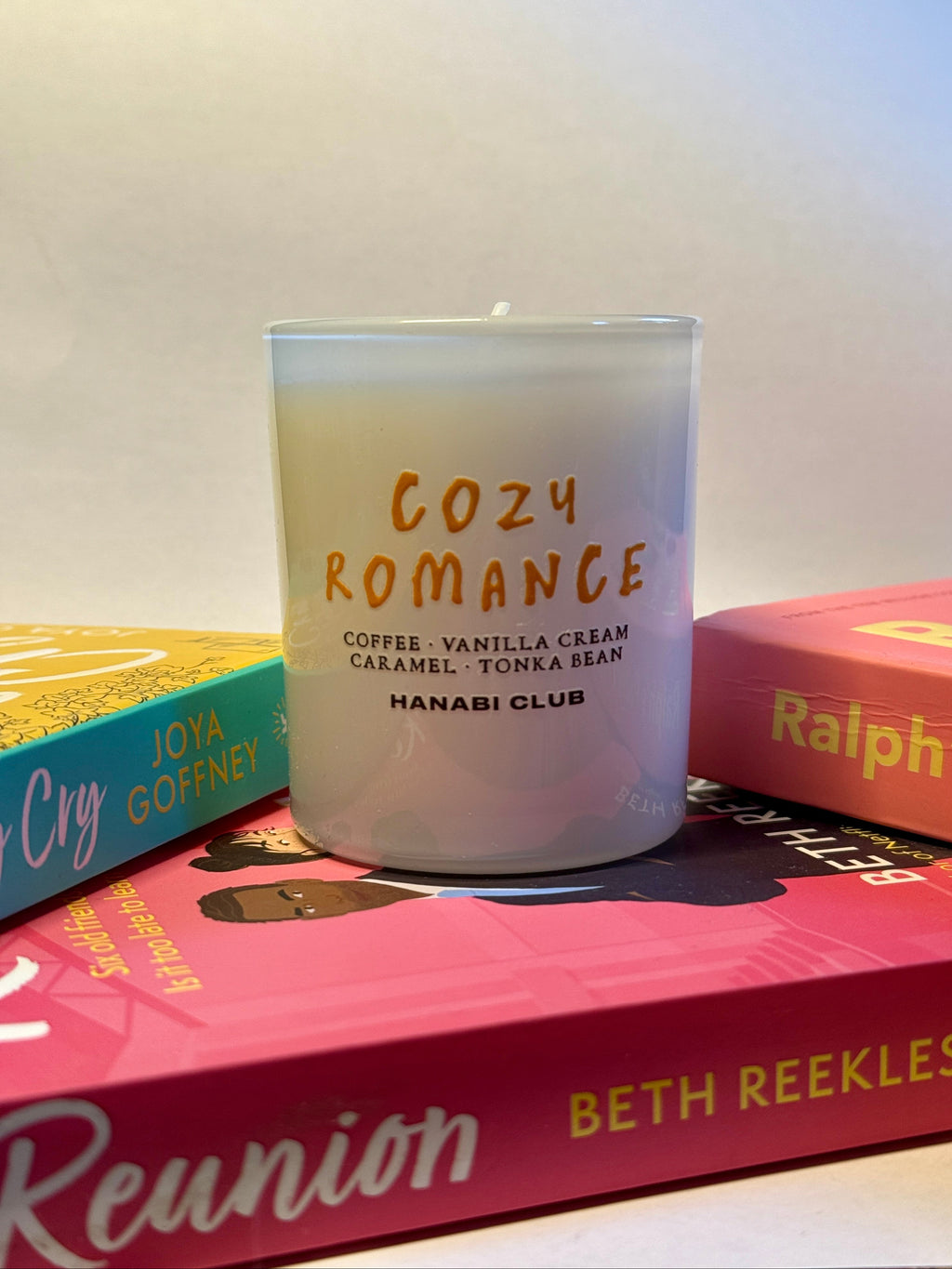A frosted glass jar candle with orange text reading 'COZY ROMANCE' and smaller scent details, styled on top of colorful book spines against a plain light background.