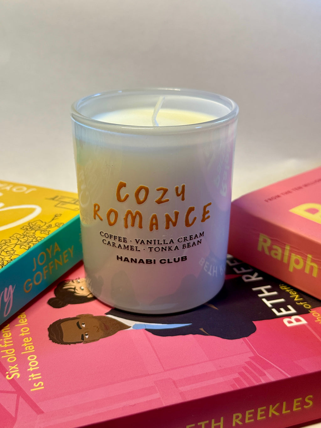 A Hanabi Club 'Cozy Romance' scented candle in a clear glass jar, styled on top of colorful books.