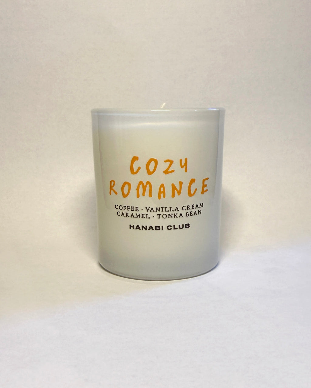 A frosted glass candle jar with orange and black text reading 'COZY ROMANCE' and 'HANABI CLUB', photographed against a plain off-white background.