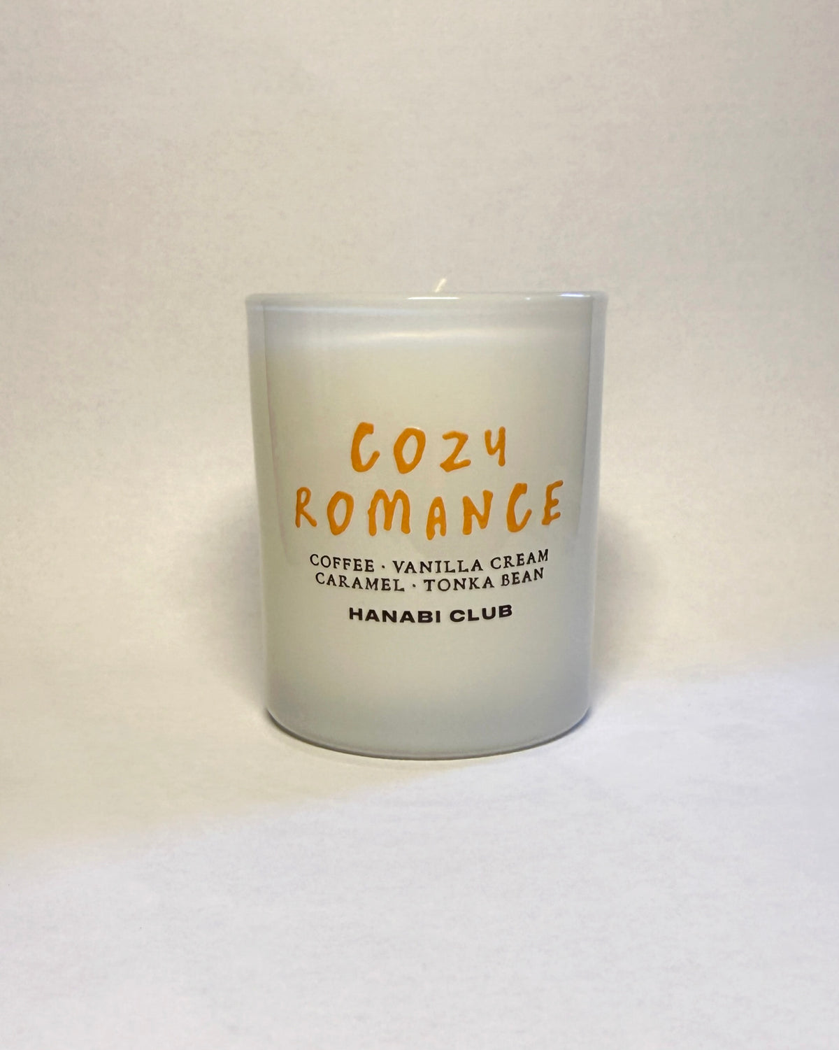 A frosted glass candle jar with orange and black text reading 'COZY ROMANCE' and 'HANABI CLUB', photographed against a plain off-white background.