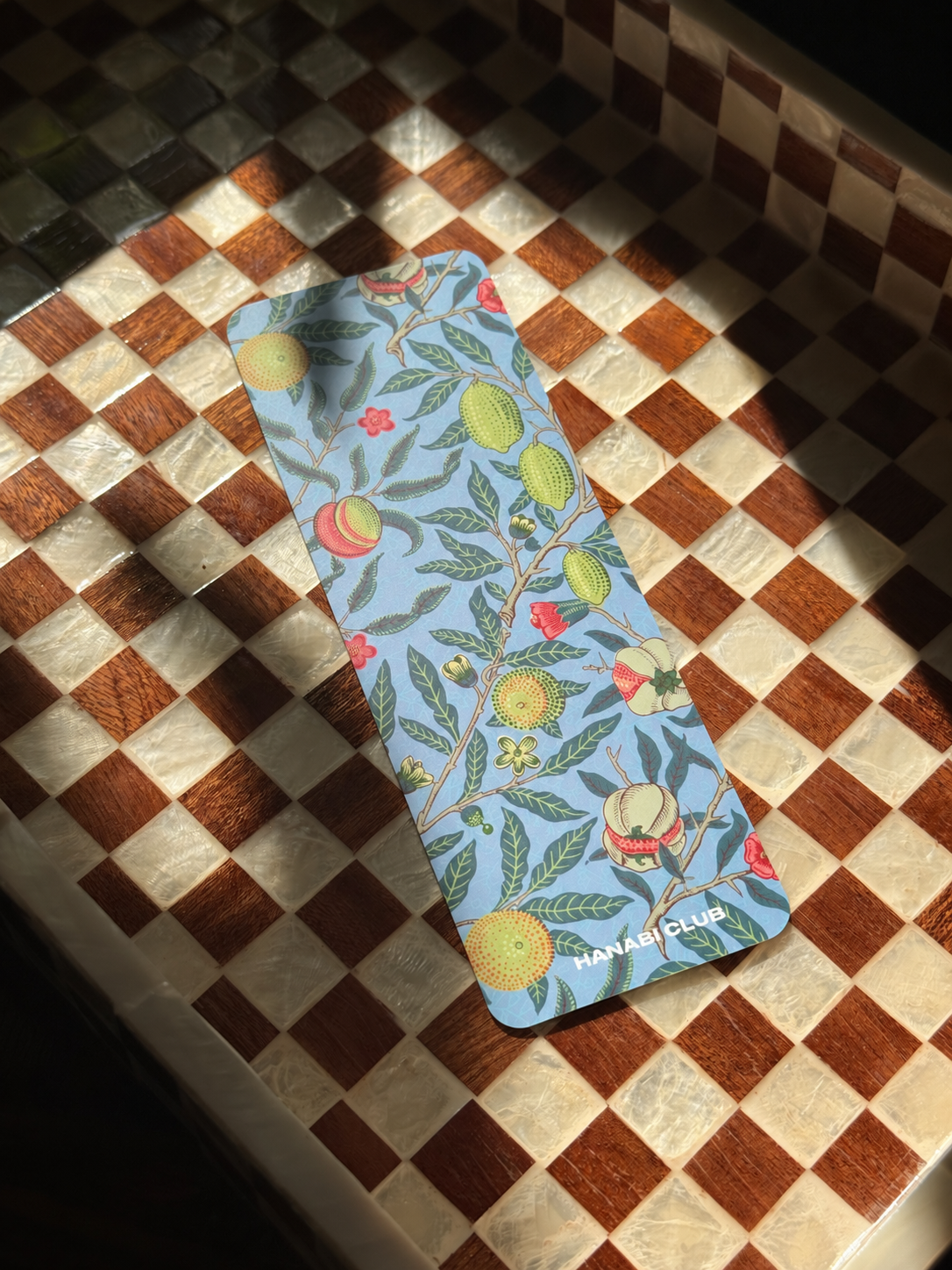 A slim rectangular card or bookmark with a light blue background and a botanical fruit print, placed diagonally on a brown and cream checkered wooden surface under direct sunlight.