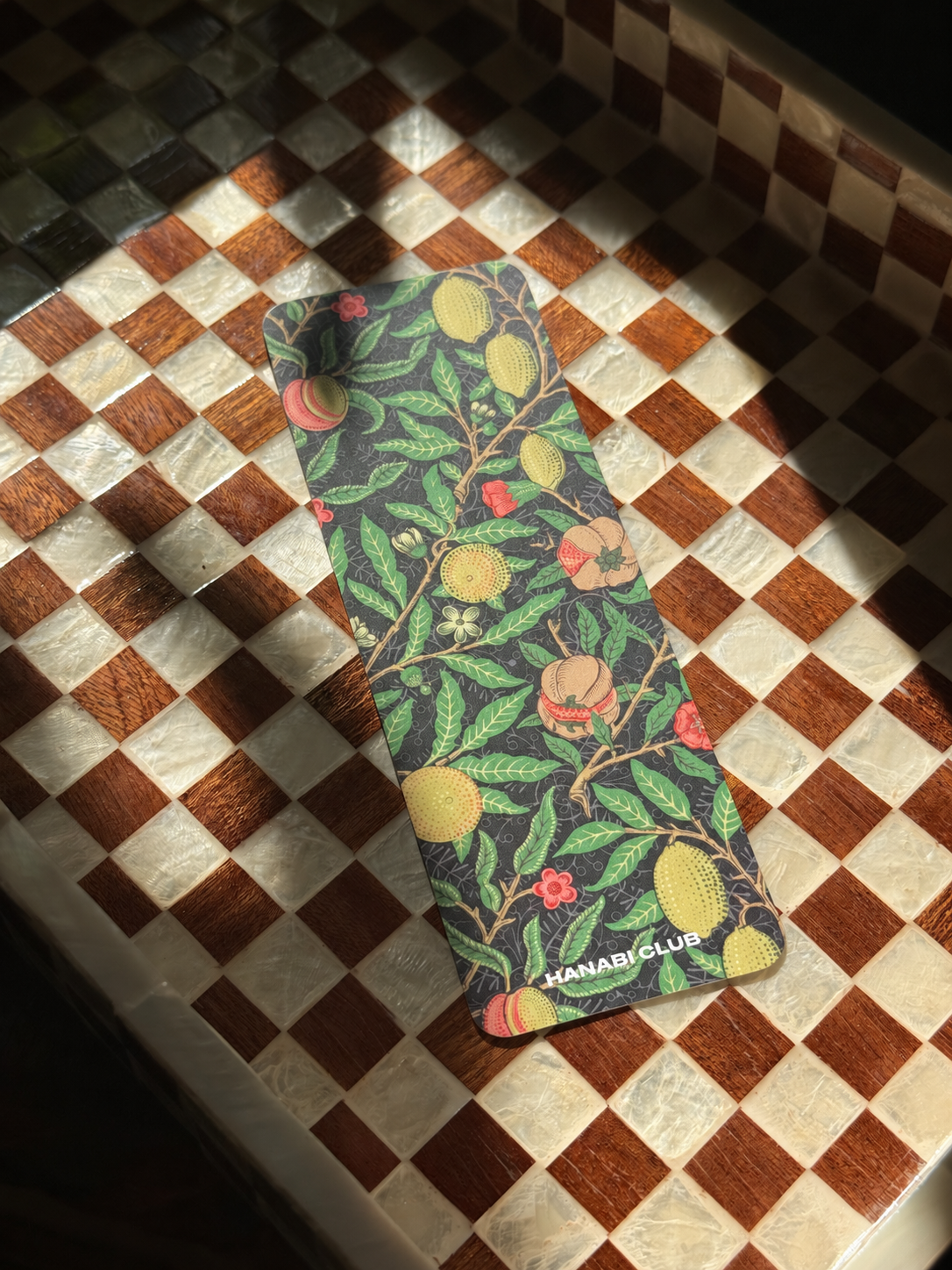 A rectangular yoga mat with a botanical pattern of green leaves, yellow peaches, and red flowers, placed on a wooden and marble checkered tray under direct sunlight. The brand name 'HANABI CLUB' is printed at the bottom.