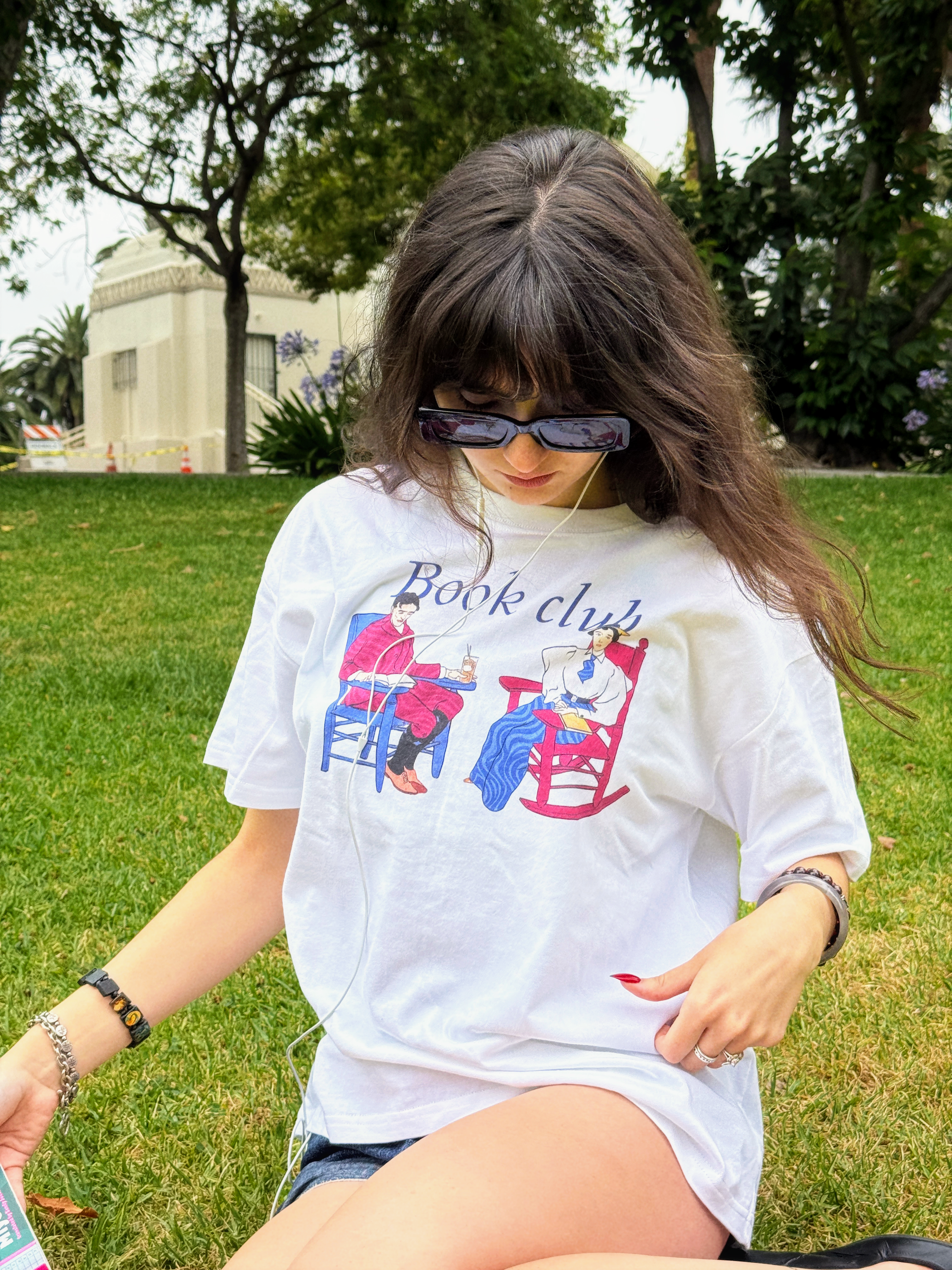 Model wearing the HANABI CLUB “Book Club Lounge” T-shirt while sitting in a park.