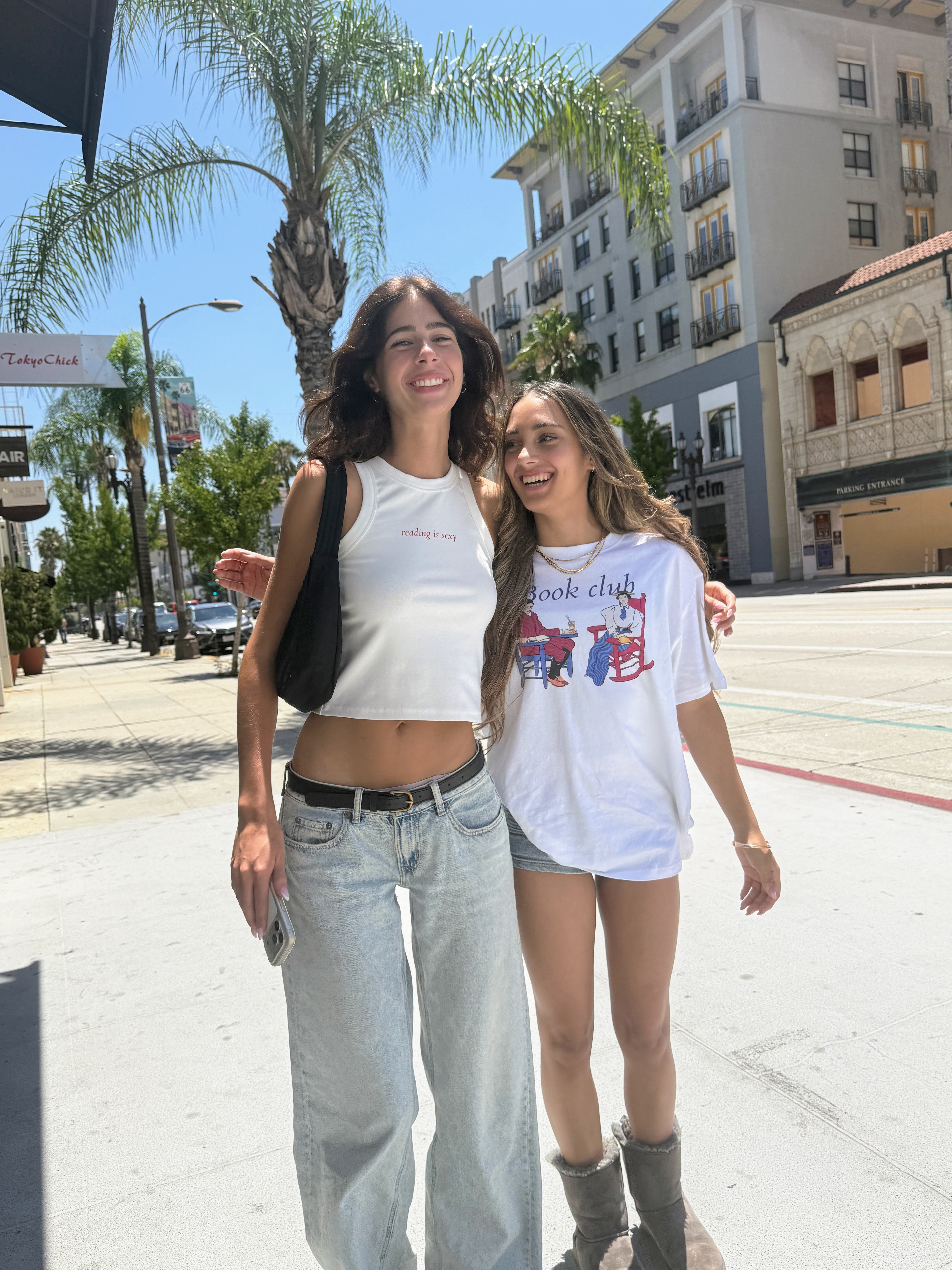Two models walking outdoors, one wearing the “Book Club Lounge” T-shirt with denim shorts.