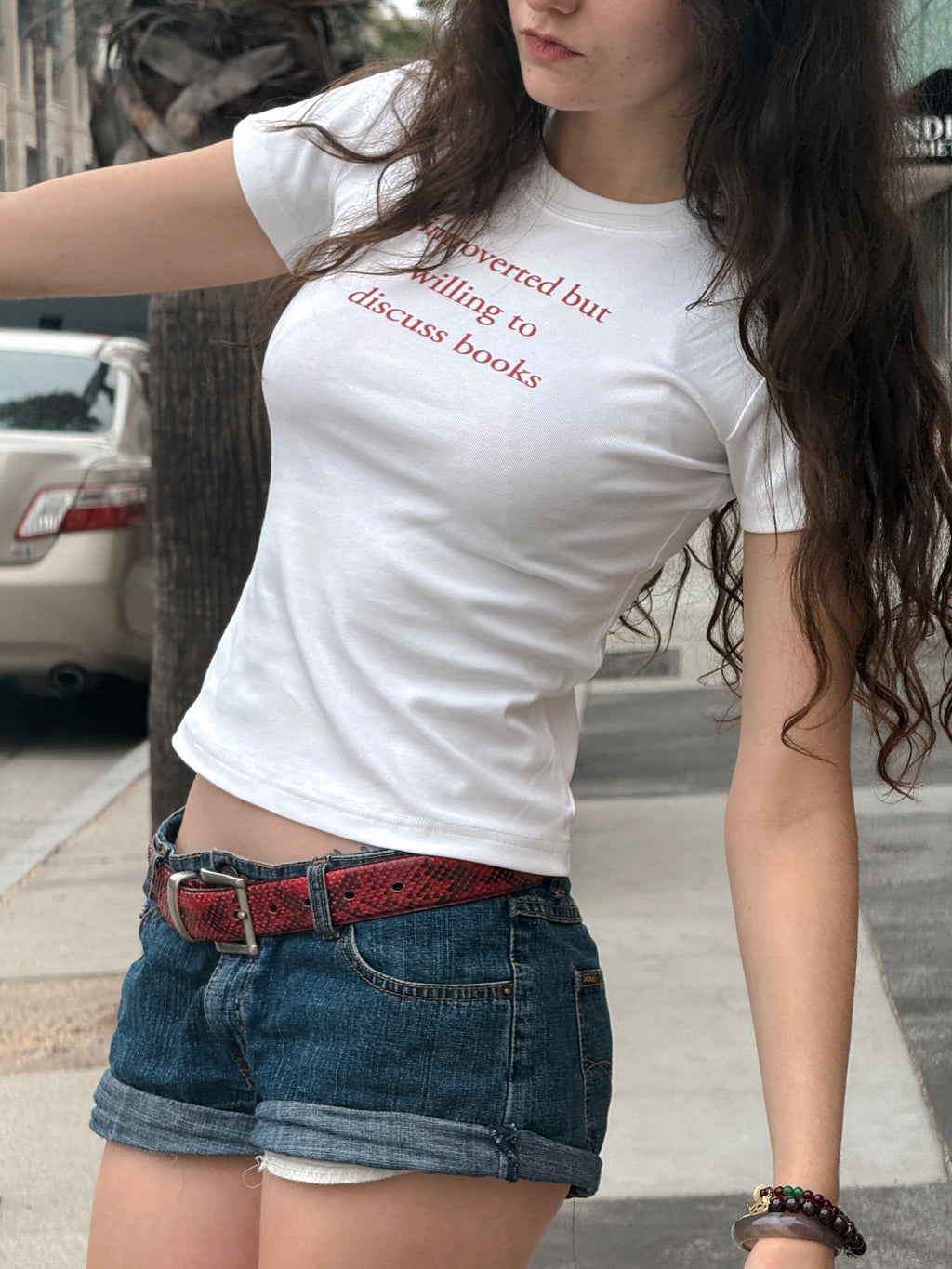 Angled view of the “Introverted but willing to discuss books” baby tee styled with denim shorts and a red belt.
