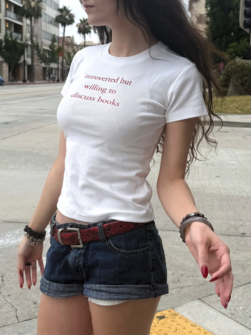 Model wearing the “Introverted but willing to discuss books” baby tee with denim shorts and a red belt outdoors.