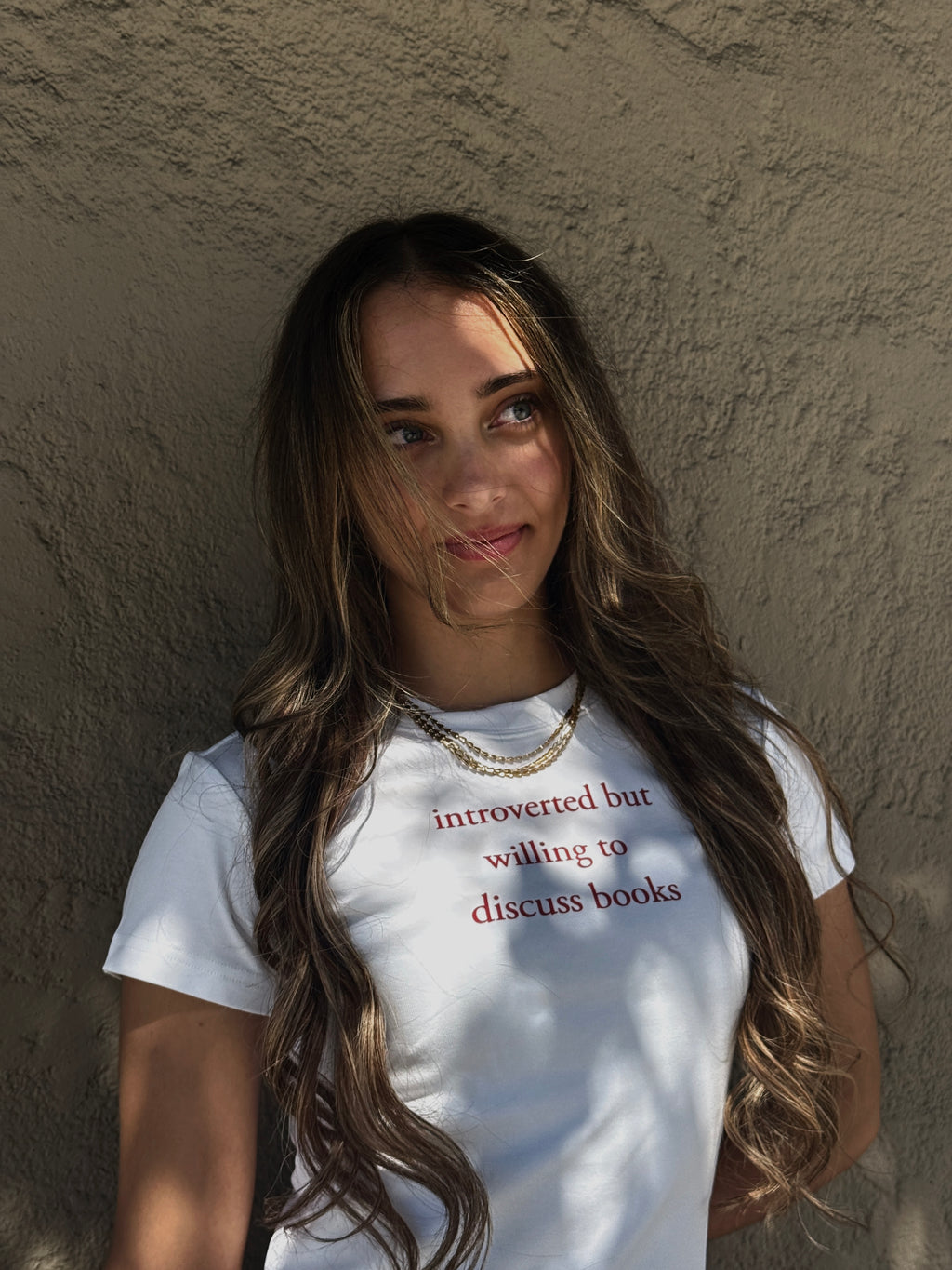 Model wearing the HANABI CLUB “Introverted but willing to discuss books” baby tee outdoors against a wall.