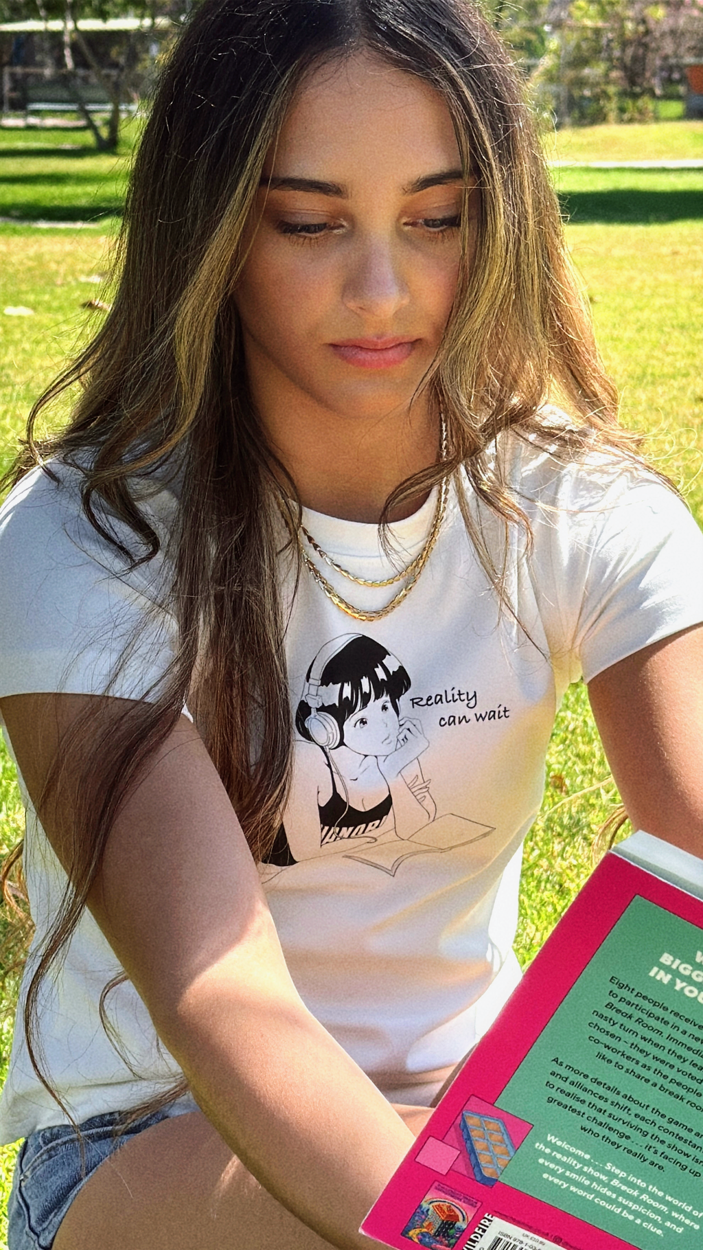 Close-up of the “Reality Can Wait” baby tee while the model reads outdoors.