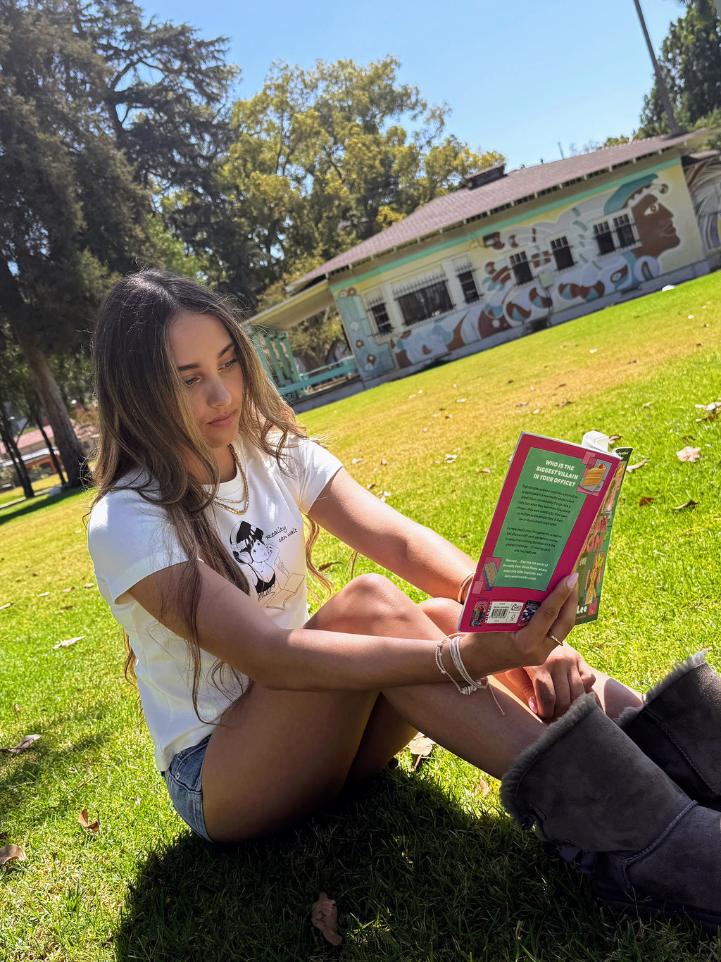 Model sitting in a park reading while wearing the “Reality Can Wait” baby tee.