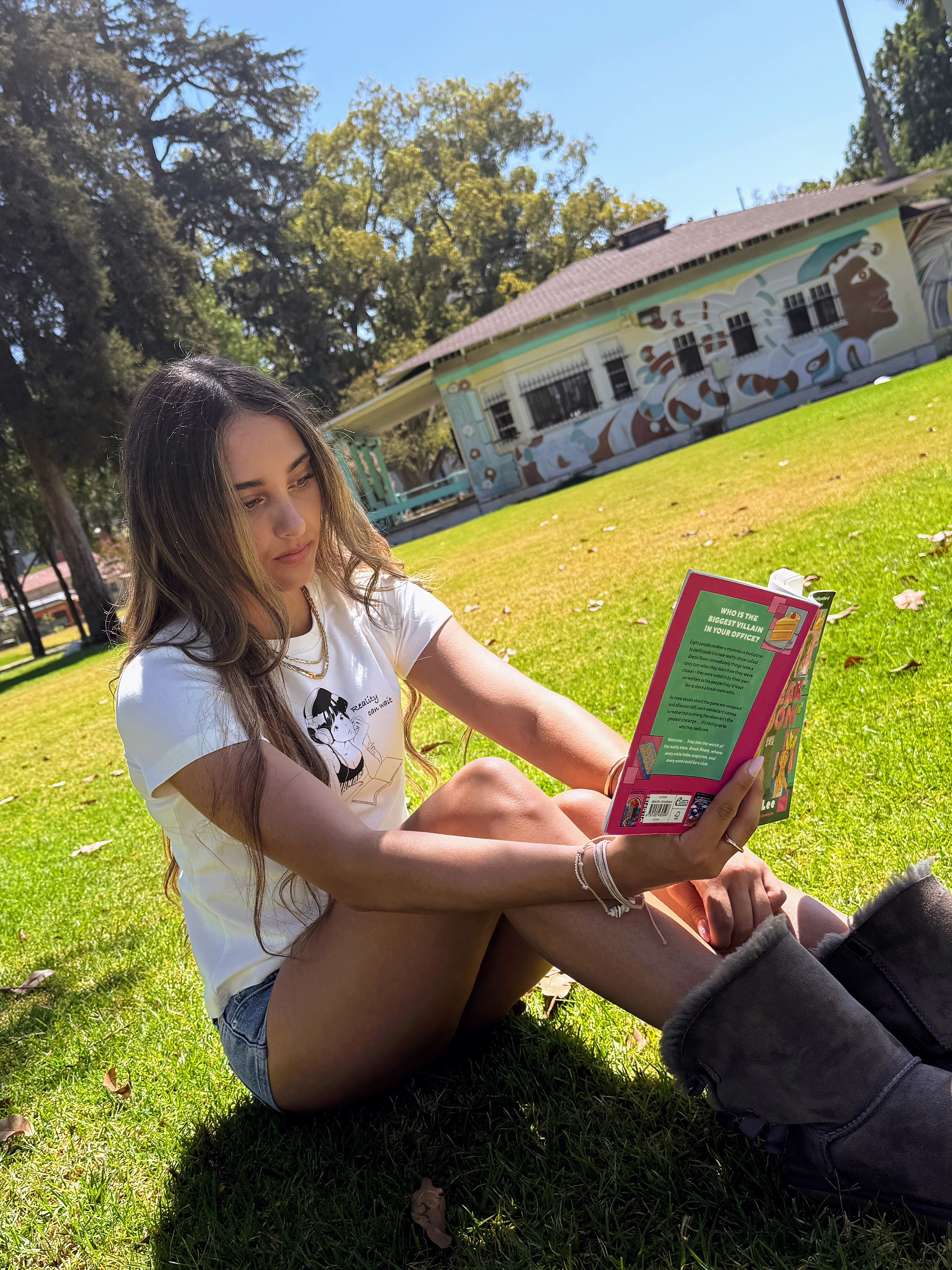 Model sitting in a park reading while wearing the “Reality Can Wait” baby tee.