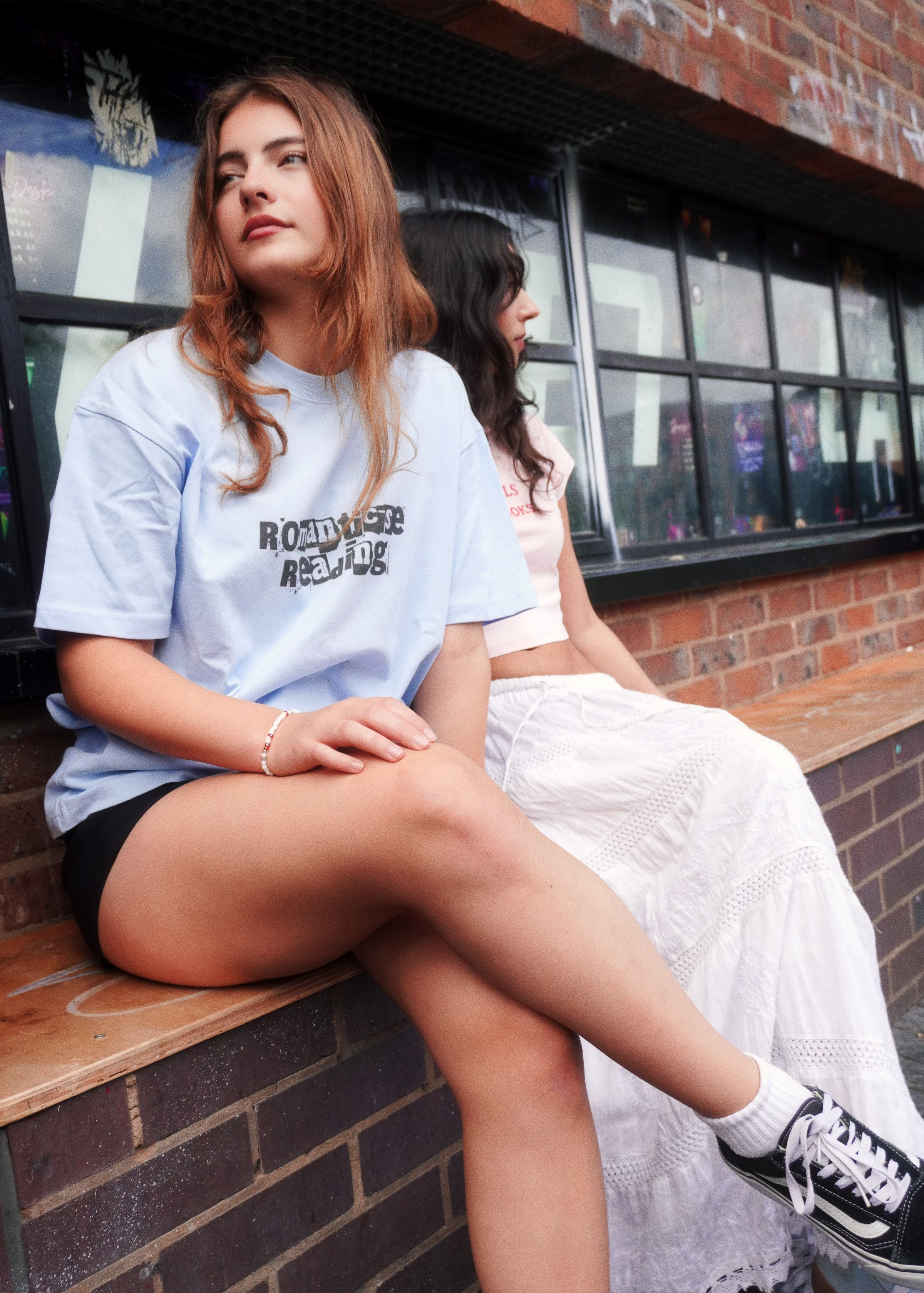 Two models sitting by a brick wall, one wearing the HANABI CLUB “Romanticise Reading” T-shirt.