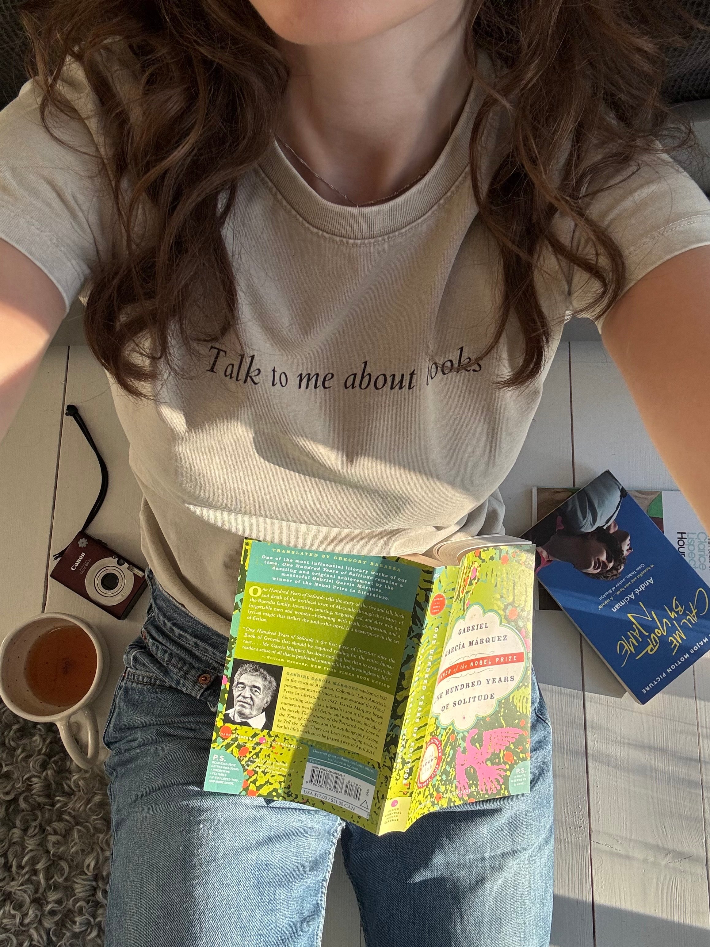 Model wearing the “Talk to me about books” ladies tee with jeans while reading indoors.