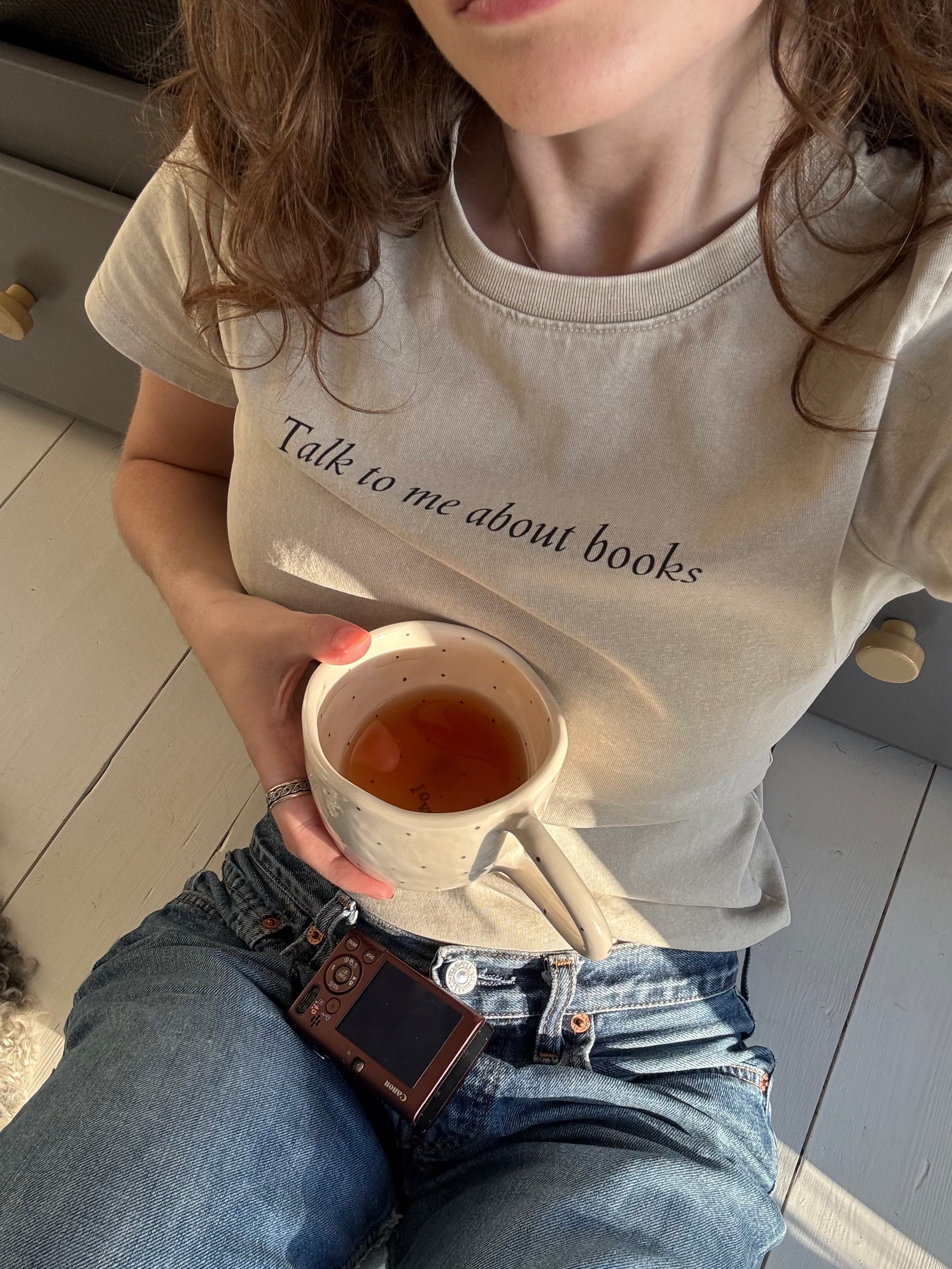 Close-up of the “Talk to me about books” ladies tee styled with jeans, a mug and a camera.