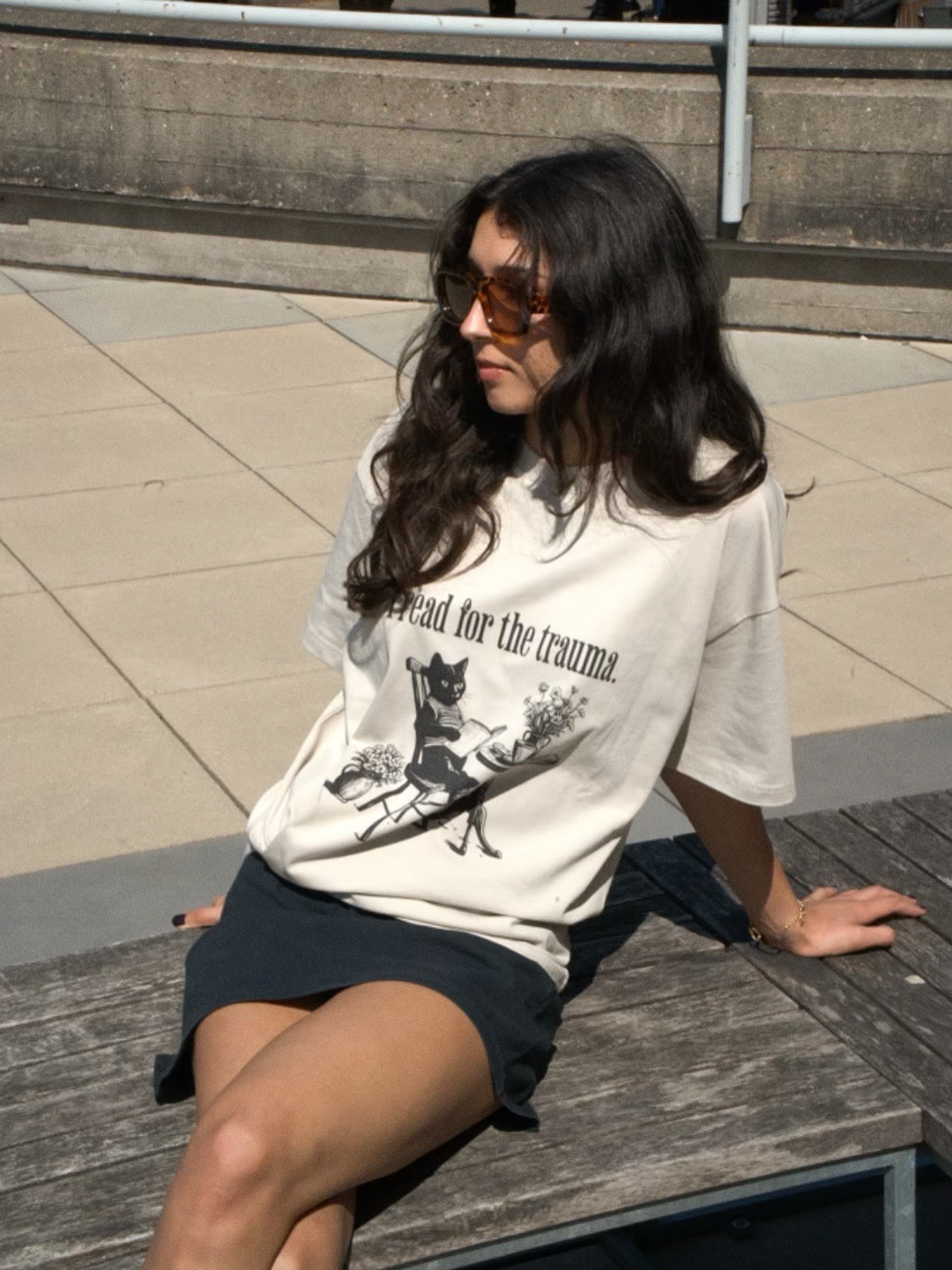 woman wearing a HANABI CLUB 'I read for the trauma' tee, whilst sitting on a bench.