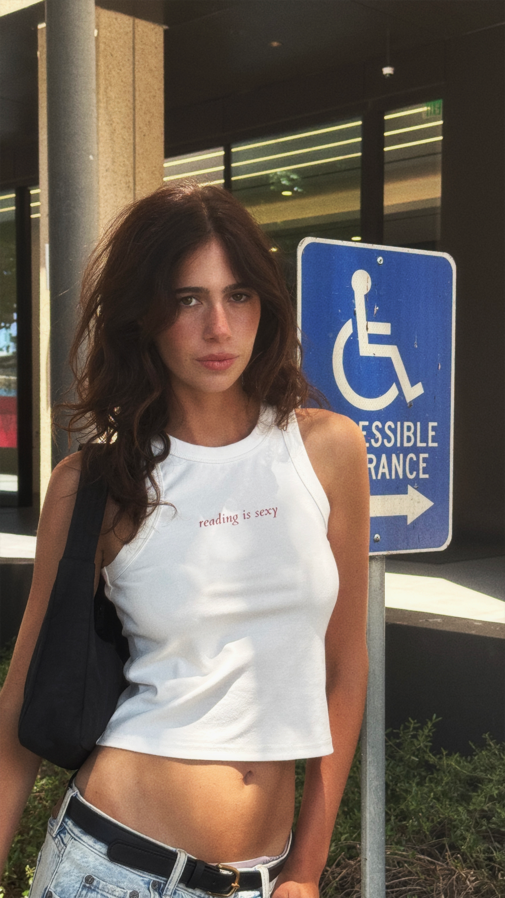 Model wearing a white “reading is sexy” tank top with light-wash jeans and a black shoulder bag outdoors.