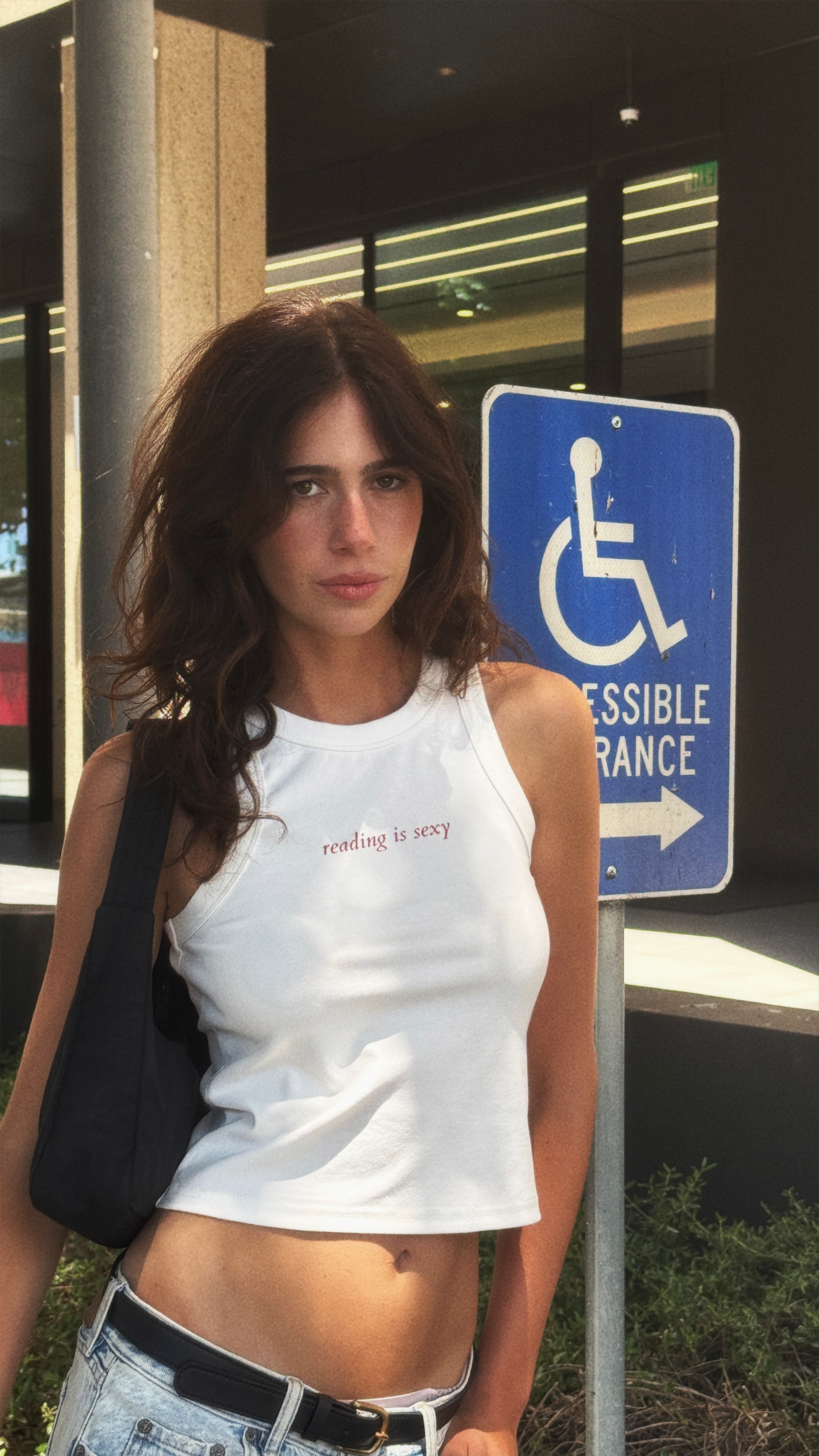 Model wearing a white “reading is sexy” tank top with light-wash jeans and a black shoulder bag outdoors.