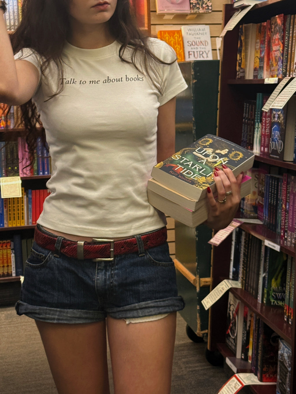 A woman in a bookshop holding a stack of books, wearing a white t-shirt with the text 'Talk to me about books' and dark denim shorts.